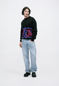 Junger Mann mit schwarzem Grafikpullover, hellblauen, lässig geschnittenen Jeans und schwarzen klobigen Schuhen, der mit einer Hand in der Tasche steht.