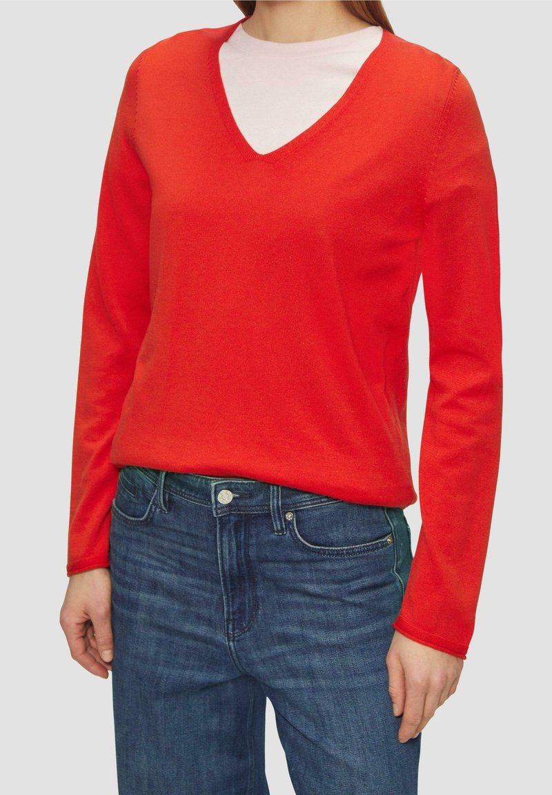 Czerwony sweter z długim rękawem i dekoltem w serek, wykonany z miękkiego materiału, noszony na jasnoróżowej bluzce z okrągłym dekoltem, zestawiony z niebieskimi jeansami. Prosty, klasyczny fason.