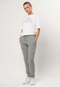 Kvinde iført hvid kortærmet skjorte med logo, grå sweatpants med snøre, hvide sneakers, stående med hænderne i lommerne mod hvid baggrund.