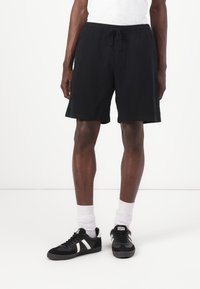Schwarze Baumwollshorts mit einem elastischen Bund und Kordelzug. Getragen mit schwarzen Sneakern mit weißen Streifen und weißen Socken.