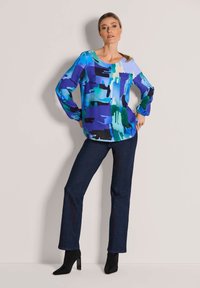 Langärmlige Bluse in blauem und grünem abstraktem Muster aus glattem Stoff, kombiniert mit dunklen Denim-Hosen und schwarzen Stiefeln mit Absatz.