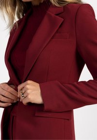 Blazer bordeaux au design ajusté, avec un revers cranté et une poche poitrine unique. Texture lisse, manches longues et silhouette cintrée.