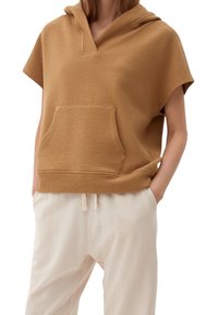 Hnědá mikina s kapucí z měkké, strukturované tkaniny, s klokaní kapsou a krátkými rukávy, kombinovaná se světle béžovými kalhotami.