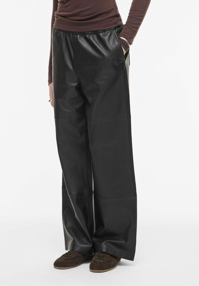 Personne portant un pantalon en cuir noir ample, un haut marron à manches longues et des chaussures marron, debout avec une main dans la poche, sur fond blanc.