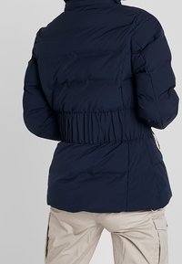 Tmavě modrá péřová bunda s elastickým pasem, měkkou texturou, vysokým límcem a prošíváním. Lehký a izolovaný design.