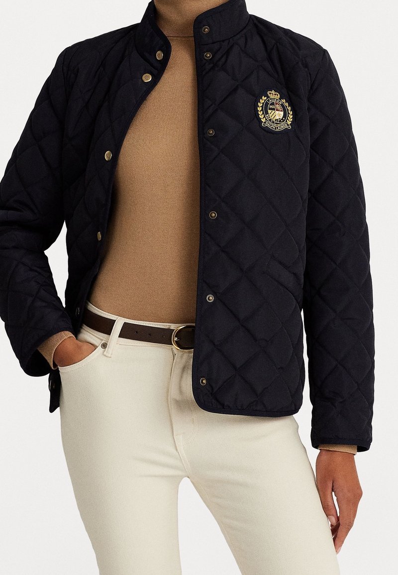 Prošívaná námořnicky modrá bunda se stojatým límcem, zlatými knoflíky a vyšívaným znakem; nošená přes béžový top a bílé džíny s hnědým opaskem.