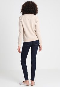 Sudadera beige con mangas raglán, puños y cinturilla de canalé, combinada con jeans oscuros ajustados y zapatos ligeros tipo slip-on, vista desde la parte posterior.