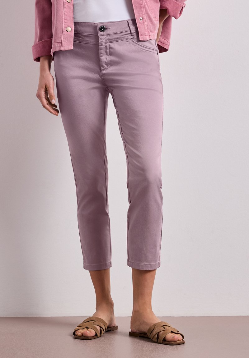 Frau trägt mauvefarbene, gekürzte Hose, eine pinke Jacke, ein weißes Top und beige gewebte Pantoletten, steht auf einem neutralen Boden.