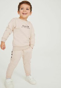 Lyserød sweatshirt og matchende joggingbukser; har et logo på brystet og sideaccenter på bukserne. Parret med hvide sneakers.
