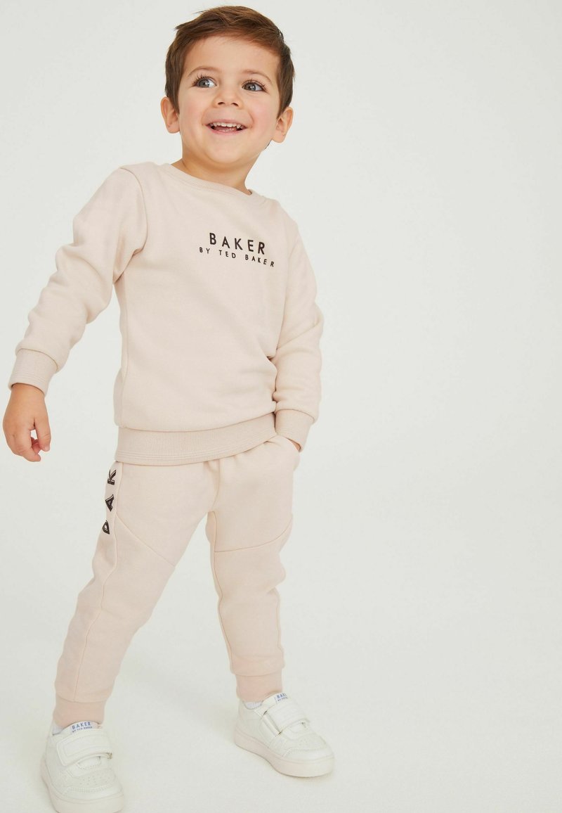 Lyserød sweatshirt og matchende joggingbukser; har et logo på brystet og sideaccenter på bukserne. Parret med hvide sneakers.