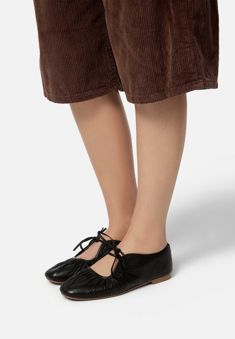 Ballerines en cuir noir avec un bout arrondi, design plissé et lacets noués. Les chaussures ont une semelle plate et un style minimaliste.