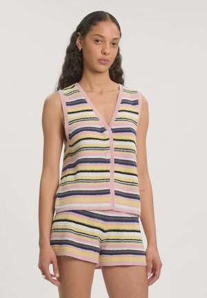 Vrouw met lang golvend haar die een mouwloos V-hals vest met knopen en bijpassende korte broek met horizontale pastelstrepen draagt.