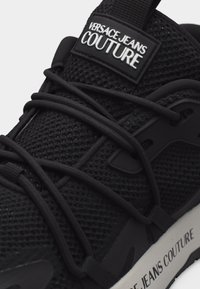 Μαύρα αθλητικά παπούτσια από διχτυωτό υλικό με μαύρα κορδόνια, ετικέτα «VERSACE JEANS COUTURE» στη γλώσσα και κείμενο στην άκρη της λευκής ενδιάμεσης σόλας.