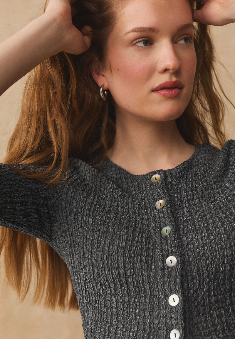 Jeune femme ajustant ses longs cheveux roux, portant de petites boucles d'oreilles créoles et un haut boutonné texturé gris charbon.