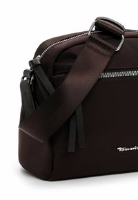 Tumši brūna taisnstūra plecu soma ar sudraba rāvējslēdzēju, plašu regulējamu siksnu un mazu priekšpuses logotipu uz balta fona.