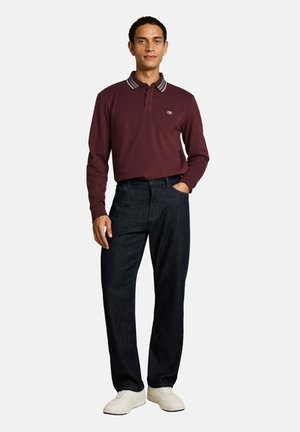 Uomo in piedi che indossa una polo a maniche lunghe rosso scuro, jeans blu scuro e scarpe da ginnastica bianche, con una mano in tasca.