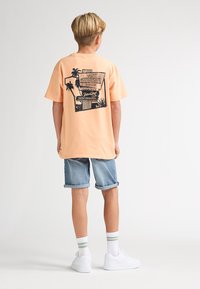 Perzikkleurig T-shirt met zwarte grafische print van palmbomen op de achterkant; gecombineerd met opgerolde spijkershorts en witte sneakers.