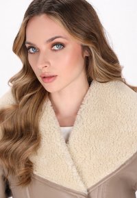 Giacca beige foderata in shearling con un ampio colletto crema, caratterizzata da una texture liscia e design angolato. Capelli che cadono in morbide onde.