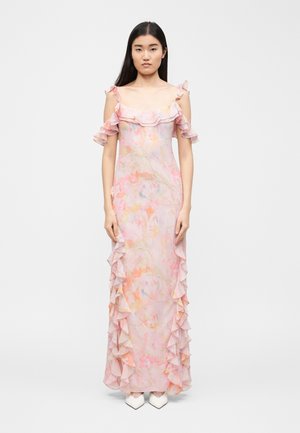 Žena v dlhých narazových šatách s pastelovým kvetinovým vzorom, s riasenými odhalenými ramenami a kaskádovitými volánmi po bokoch, stojaca na bielom pozadí.