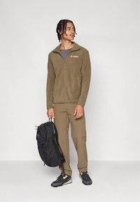 Õlivärvi fliisjakk esi lukuga ja logoga, kombineeritud beežide pükstega ning musta seljakotiga. Jalatsid on musta värvi hallide aktsentidega.