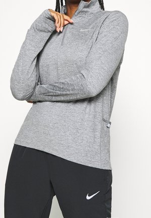 Osoba nosząca szary długi rękaw top Nike z zamkiem oraz czarne spodnie Nike, z rękami skrzyżowanymi i jedną ręką blisko twarzy.