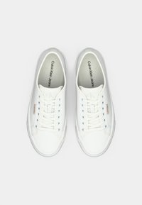 Ein Paar weiße Calvin Klein Jeans Sneaker mit Schnürsenkeln, grauen Sohlen und kleinen roten Logopatches auf einem hellgrauen Hintergrund.