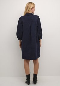 Robe bleu marine, longueur au genou, avec un col, des manches bouffantes et un pli au dos. Fabriquée en tissu texturé, avec des détails de boutons aux poignets.