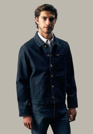 Junger Mann trägt dunkelblaue Jeansjacke und Jeans, steht vor einem einfarbigen beigen Hintergrund und schaut mit neutralem Gesichtsausdruck geradeaus.