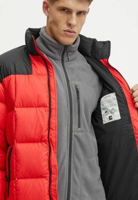 Giacca a vento rossa e nera con collo alto, indossata sopra un pile grigio. Presenta tasche con zip e un'etichetta di cura all'interno.