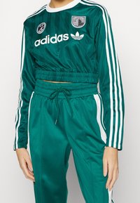 Teal adidas crop-top med långa ärmar och elastisk midja. Har vit logotyp och ränder. Matchas med matchande avslappnade träningbyxor.