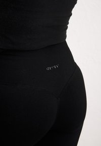 Gros plan d'une personne portant des leggings noirs taille haute avec une ceinture côtelée et un petit logo au dos.