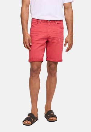 Hombre con pantalones cortos rojos remangados, camiseta blanca y sandalias negras, de pie contra un fondo liso y claro.