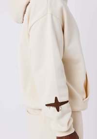 Felpa crema con vestibilità rilassata, caratterizzata da un ricamo a forma di stella marrone sul polsino. Tessuto morbido con una superficie liscia.
