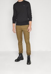 Pull noir en maille avec poignets et ourlet côtelés, associé à un pantalon marron ajusté et des bottes noires à lacets. Design simple avec des couleurs neutres.