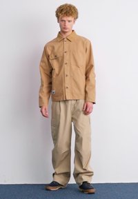 En beige knapp-up jakke med to brystlommer, kombinert med løse beige cargo-bukser og mørke sko, mot en ensfarget bakgrunn.