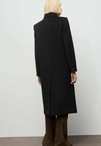 Abrigo de lana negro con una silueta larga y recta, cuello con muesca y puños abotonados, mostrado desde atrás sobre un fondo claro.