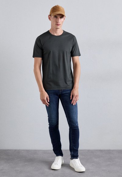 Homme portant une casquette beige, un t-shirt gris foncé, un jean skinny bleu et des baskets blanches, se tenant contre un fond gris clair uni.
