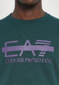 T-shirt verde de mangas curtas com um grande logótipo gráfico roxo "EA" e o texto "EMPORIO ARMANI" abaixo. Tecido de algodão suave, decote redondo.