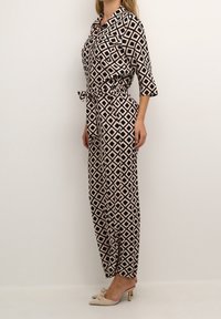 Combinaison à motifs avec un design géométrique en noir, beige et rose. Comprend une taille cintrée, des manches trois-quarts et des poches.