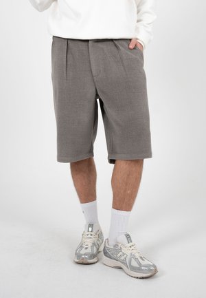 Mann iført grå shorts til knéet, hvite crew-sokker og sølv- og hvite sportssko, stående med en hånd i lomma.