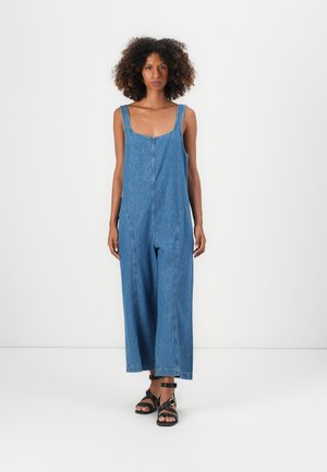 Denim overall jumpsuit with wide legs, square neckline, and side pockets. Worn with black strappy sandals, against a plain white background.