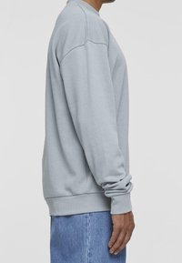 Felpa grigia chiara con collo rotondo, polsini e orlo a coste. Presenta una vestibilità rilassata e una texture morbida, indossata sopra jeans blu.