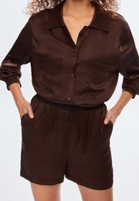 Bruin satijnen tweedelig set bestaande uit een overhemd met knopen en twee borstzakken, en shorts met elastische tailleband. Zachte textuur, ontspannen pasvorm.