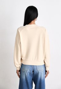 Sudadera color crema con un ajuste relajado, puños y dobladillo acanalados. Se lleva con vaqueros de lavado claro. Textura suave y lisa.