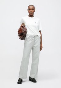 Γυναίκα με λευκό μπλουζάκι, γκρι φόρμες, μαύρες λοφίδια, κρατώντας μεγάλη καφέ δερμάτινη τσάντα μπροστά σε λευκό φόντο.