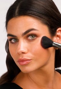 Brocha de maquillaje aplicando rubor en una mejilla, con cerdas suaves y negras y un casquillo metálico plateado. La persona tiene la piel suave y luminosa.