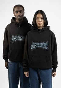 Black hooded sweatshirts with front kangaroo pockets, featuring a blue flame text graphic. Models wear loose-fitting blue denim jeans.