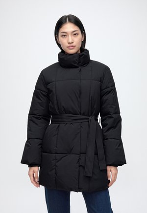 Une femme présente un manteau noir matelassé, ceinturé et à col montant, porté sur un jean bleu, devant un fond clair et uni.
