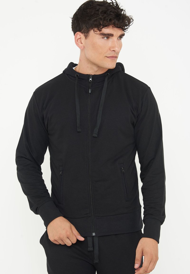 Felpa nera con zip realizzata in tessuto liscio, dotata di colletto alto, cappuccio con lacci, tasche laterali e polsini a coste.
