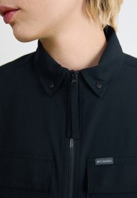 Veste noire avec une fermeture éclair à l'avant, des boutons-pression et deux poches poitrine. Le matériau semble texturé, avec une étiquette de logo sur la poche de gauche.